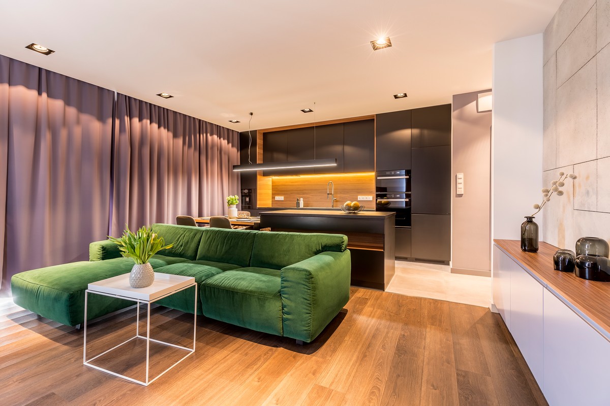 Open space apartment interior with white table near green corner sofa in front of a black, glossy kitchen with dining table